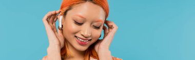 happy face, radiant smile, young asian woman fixing dyed red hair while listening music in wireless earphone on blue background, summer vibes, youth culture, banner clipart
