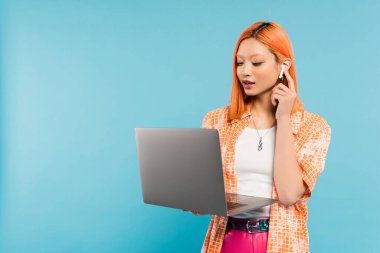 young and trendy asian woman in orange shirt, with dyed hair, adjusting wireless earphone and looking at laptop when standing on blue background, youth fashion, freelance lifestyle, generation z clipart