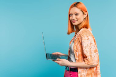 happy face, optimistic emotion, youthful asian woman with colored red hair, in orange shirt holding laptop and looking at camera on blue background, freelance lifestyle, generation z clipart