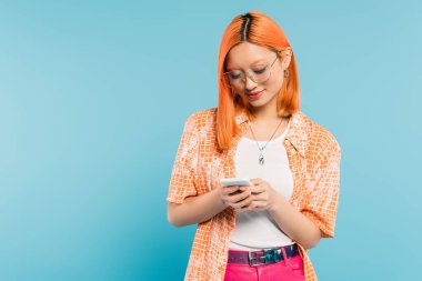 digital lifestyle, positive emotion, smiling asian woman with dyed red hair, in stylish eyeglasses and orange shirt networking on mobile phone on blue background, generation z, summer vibes clipart