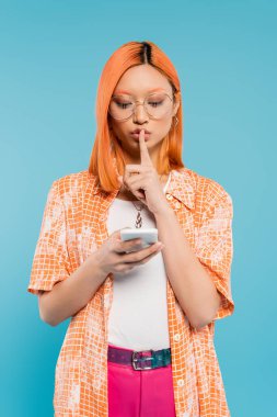 Gizlilik, gizlilik, gözlüklü ciddi bir Asyalı kadın mavi arka plandaki akıllı telefondan telefon görüşmesinde sessizlik işareti gösteriyor, kırmızı renk saç, turuncu gömlek, modern moda, z nesli.