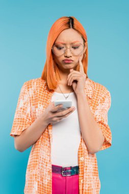 negative emotion, bad mood, displeased asian woman touching cheek while looking at smartphone on blue background, trendy eyeglasses, red colored hair, orange shirt, youthful fashion clipart