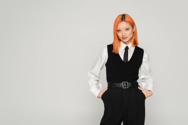 self-assured asian woman with colored red hair posing with hands in pockets in white shirt, black tie, vest and pants when looking at camera on grey background, business casual fashion clipart