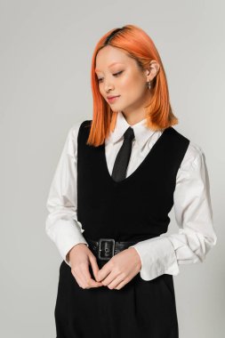 Çekici ve pozitif Asyalı kadın, gri arka planda duran ve gülümseyen kızıl saçlı, beyaz gömlekli, siyah kravatlı ve yelekli iş kadını, günlük moda, Z jenerasyonu, modern yaşam tarzı.