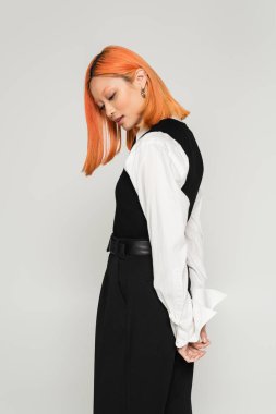 Gri arka planda elleri arkadan poz veren, etkileyici ve genç Asyalı kadın, boyalı kızıl saç, beyaz gömlek, yelek ve pantolon, iş gündelik moda, modern yaşam tarzı.