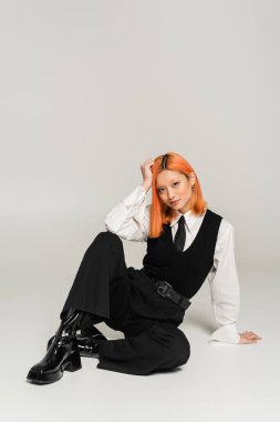 young asian woman with colored red hair, in black and white clothes sitting and looking at camera on grey background, white shirt, black pants and vest, business casual fashion, full length clipart