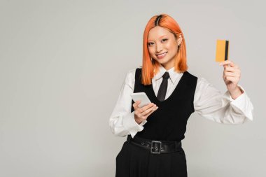 Pozitif duygu, boyalı kızıl saçlı neşeli Asyalı kadın akıllı telefon tutuyor, kameraya bakıyor ve gri arka planda kredi kartı gösteriyor, iş kıyafeti, beyaz gömlek, siyah kravat ve yelek, genel z