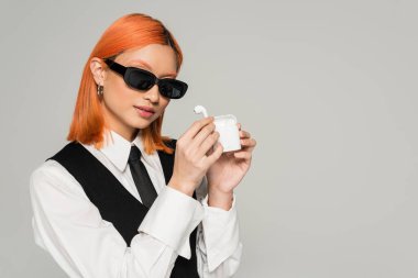 Modern müzik aşığı, koyu kırmızı saçlı, koyu güneş gözlüklü, beyaz gömlekli, siyah kravatlı ve yelekli gri arka planda kablosuz kulaklıklı Asyalı kadın.