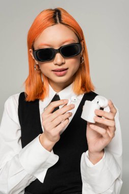 red haired and youthful asian woman in dark stylish sunglasses holding case with wireless earphones on grey background, white shirt, black tie and vest, business casual fashion, generation z clipart