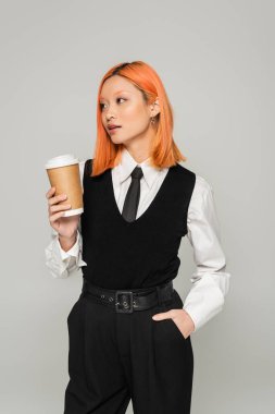 Genç ve modaya uygun Asyalı kadın boyalı kızıl saçlı ve kahve sipariş eden elini cebine atmış gri arka plan, beyaz gömlek, siyah kravat, yelek ve pantolon, iş modası.