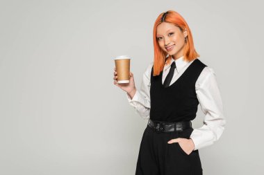 Olumlu duygular, boyalı kızıl saçlı, parlak gülüşlü, elinde sıcak içecek olan, gri arka planda kameraya bakan, siyah beyaz iş kıyafetleri, modern moda...