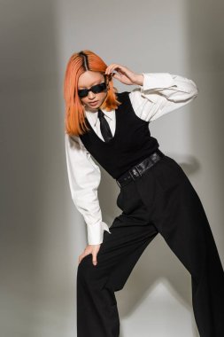 Koyu renk güneş gözlüklü modern Asyalı kadının moda çekimi. Gri gölgeli arka planda siyah ve beyaz kıyafetler. Renkli kızıl saç, beyaz gömlek, siyah kravat, yelek ve pantolon.