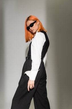 Moda fotoğrafçılığı, renkli kızıl saçlı genç Asyalı kadın. Gri gölgeli arka planda kameraya bakıyor. Koyu güneş gözlüğü, beyaz gömlek, siyah yelek, modern moda çekimi.