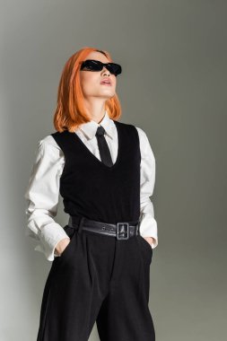 Koyu renk güneş gözlüklü, kendine güvenen Asyalı bir kadın elleri ceplerinde ve gri gölgeli arka planda, iş gündelik, beyaz gömlek, siyah kravat, yelek ve pantolon, modern moda