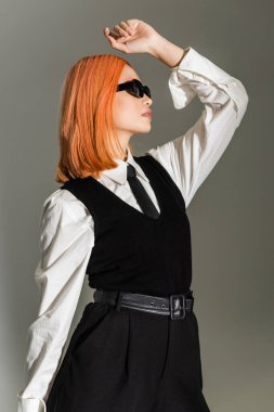 Ele avuca sığmayan gri gölgeli arka planda poz veren etkileyici ve stil sahibi Asyalı kadının yan görünüşü, boyalı kırmızı saç, koyu güneş gözlüğü, beyaz gömlek, kravat ve yelek, iş modası