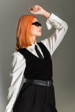 side view of expressive and stylish asian woman posing with hand above head on grey shaded background, dyed red hair, dark sunglasses, white shirt, tie and vest, business casual fashion clipart