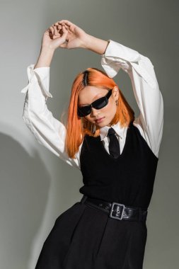 expressive asian fashion model with dyed red hair, in dark sunglasses, white shirt, black tie, pants and vest posing with hands above head on grey shaded background, business fashion photography clipart