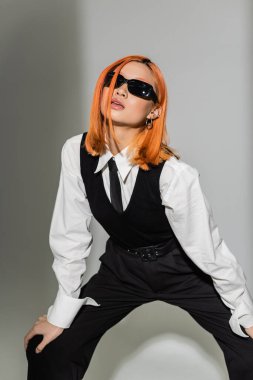 Koyu güneş gözlüklü, genç, şehvetli ve çekici Asyalı kadın, beyaz gömlek, siyah kravat, yelek ve pantolon, gri gölgeli arka plan, iş modası fotoğrafçılığı.