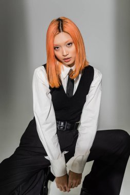 young and expressive asian woman in black and white business casual clothes looking at camera on grey shaded background, colored red hair, white shirt, black tie and pants, modern fashion clipart