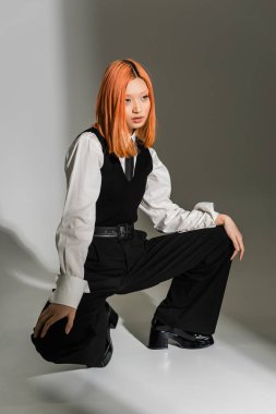 Kırmızı saçlı ve kendine güvenen Asyalı kadın şık iş kıyafetleri içinde gri renkli arka plan, beyaz gömlek, siyah yelek ve pantolon, modern moda, z nesline bakıyor ve poz veriyor.