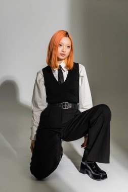 full length of youthful and stylish asian woman in a white shirt, black vest and pants, with colored red hair posing and looking away on grey shaded background, fashion shoot, generation z clipart