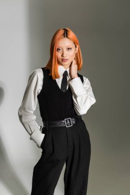 business casual fashion, young asian woman with colored red hair, in white shirt, black vest and pants posing with hand in pocket while looking at camera on grey shaded background, fashion shoot clipart