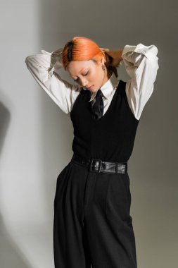 fashionable asian woman with dyed red hair, in black and white business casual clothes posing with hands behind head on grey shaded background, youthful style, generation z, youth culture clipart