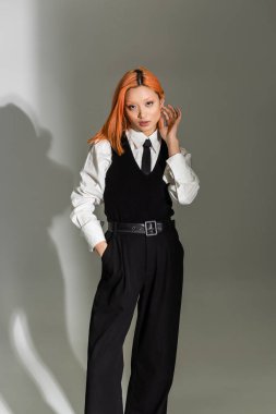 Moda ve kendinden emin Asyalı kadın kırmızı renkli saçlara dokunuyor, el ele tutuşuyor ve gri gölgeli arka planda kameraya bakıyor, iş kıyafeti, beyaz gömlek, siyah kravat, gömlek ve pantolon.