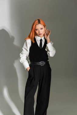 trendy and confident asian woman touching red colored hair, holding hand in pocket and looking at camera on grey shaded background, business casual, white shirt, black tie, shirt and pants clipart