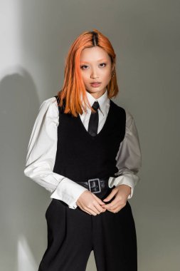 charming and red haired asian woman in white shirt, black tie, vest and pants looking at camera on grey shaded background, business casual fashion, generation z lifestyle clipart