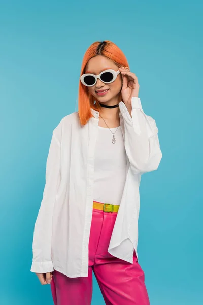 young culture, cheerful asian woman with dyed hair standing in casual attire and sunglasses, smiling on blue background, white shirt, choker necklace, red hair, generation z