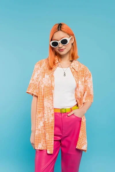 casual attire, smiling asian woman with dyed hair and sunglasses standing with hand in pocket on vibrant blue background, orange shirt, red hair, modern fashion, generation z