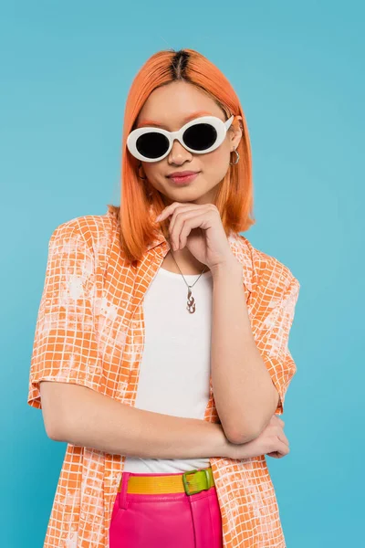 generation z, young asian woman with dyed hair standing in casual attire and sunglasses, looking at camera on vibrant blue background, orange shirt, red hair, personal style