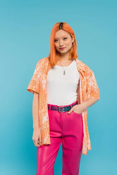 casual attire, young asian woman with dyed red hair standing with hand in pocket of pink pants on vibrant blue background, orange shirt, personal style, confidence, generation z