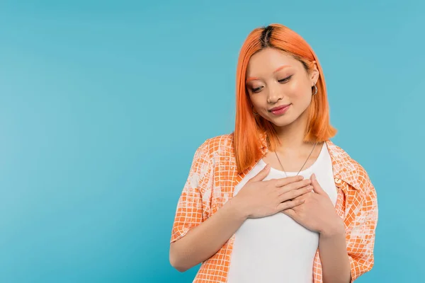 grateful, young asian woman with dyed red hair smiling and holding hands near chest on vibrant blue background, pleased, generation z, casual attire, looking away, young culture
