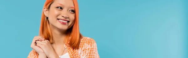 stock image positivity, young asian woman with dyed hair standing in orange shirt and posing with clenched hands on blue background, looking away, joyful, adorable, generation z, modern style, banner 