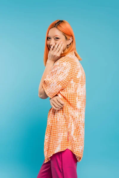 stock image positivity, shy and young asian woman with dyed hair standing in orange shirt and covering mouth with hand while smiling on blue background, looking at camera, joyful, happiness, generation z 