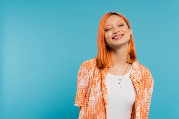 joyful face, radiant smile, young asian woman with dyed hair standing with closed eyes in orange shirt and smiling on blue background, casual attire, happiness, freedom, cheerful attitude