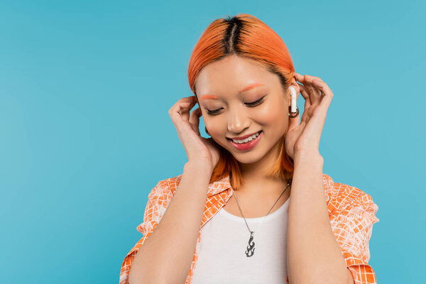 young and happy asian woman with colored red hair listening music in wireless earphone, fixing red colored hair and smiling on blue background, generation z, summer vibes