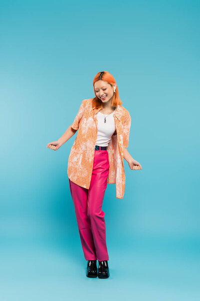 летний отдых, любитель музыки, восторженная азиатская женщина в красочной одежде и беспроводных наушниках на голубом фоне, окрашенные волосы, оранжевая рубашка, розовые брюки, ген z моды