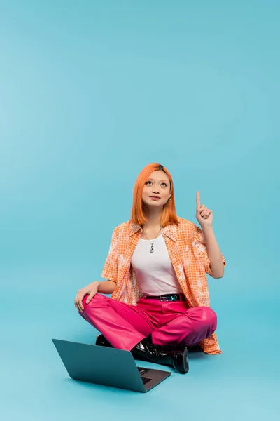 stock image creative thinking, idea gesture, solution, redhead asian woman in orange shirt sitting with crossed legs near laptop, looking up and pointing with finger on blue background, freelance lifestyle