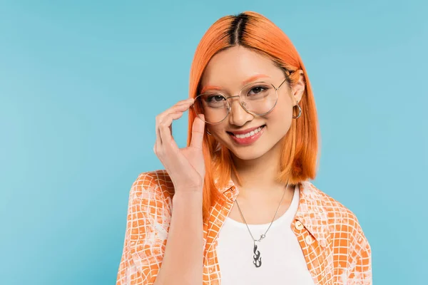 radian smile, positive emotion, happy asian woman with dyed red hair, in orange shirt adjusting eyeglasses with stylish frame and looking at camera on blue background, youthful fashion
