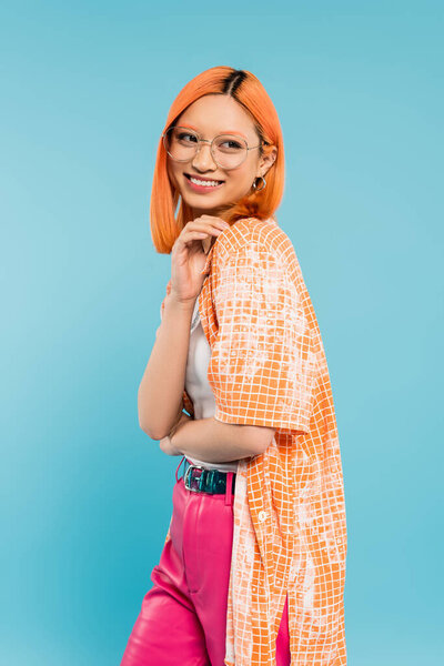 позитивные эмоции, радостная и молодая азиатская женщина с сияющей улыбкой стоя и глядя в сторону на синий фон, стильные очки, оранжевая рубашка, современная мода, поколение z