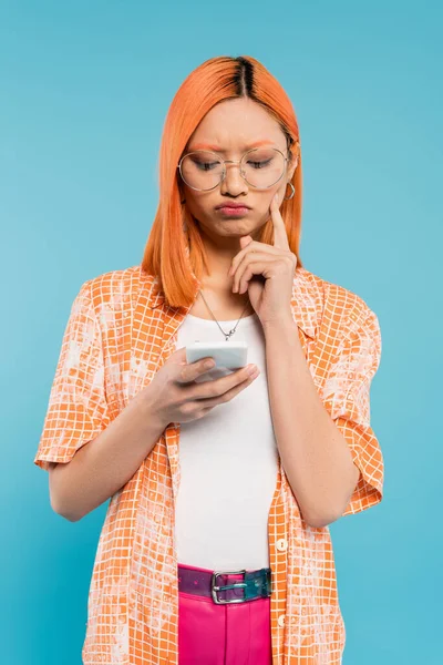 negative emotion, bad mood, displeased asian woman touching cheek while looking at smartphone on blue background, trendy eyeglasses, red colored hair, orange shirt, youthful fashion