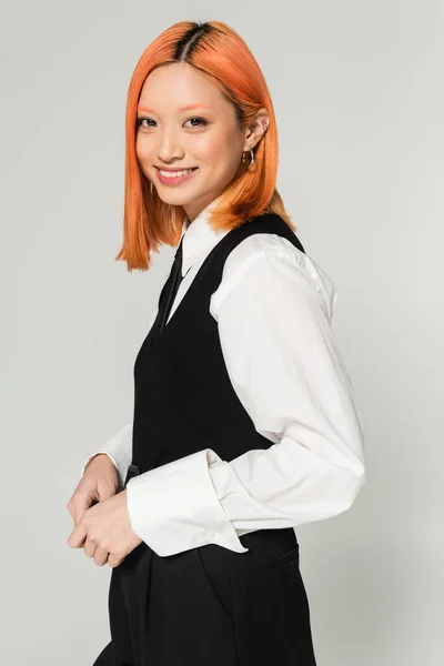 stock image positive emotion, carefree asian woman with radiant smile looking at camera on grey background, dyed red hair, white shirt, black vest, business casual style, modern fashion, generation z