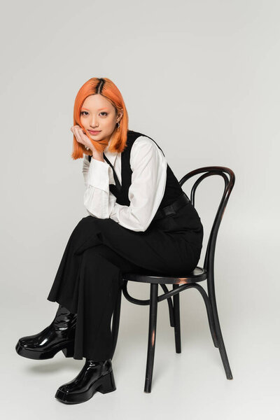 positive emotion, charming asian woman with happy face and dyed colored hair looking ta camera on chair on grey background, white shirt, black vest and pants, business fashion photography