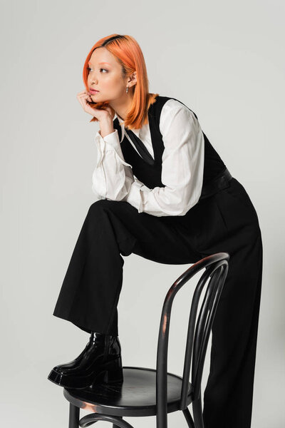 stylish and dreamy asian woman with colored red hair stepping on chair and looking away on grey background, white shirt, black vest and pants, business fashion photography, generation z