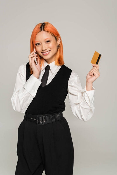 online shopping, excitement, red haired asian woman with credit card making order on smartphone on grey background, business casual style, white shirt, black vest and tie, generation z