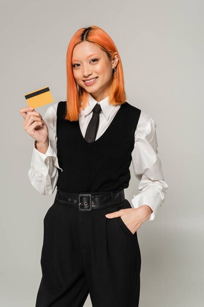 радостные эмоции, молодая и веселая азиатская женщина, стоящая с рукой в кармане и показывая кредитную карту на сером фоне, черно-белая одежда, деловые случайные, поколение z образ жизни