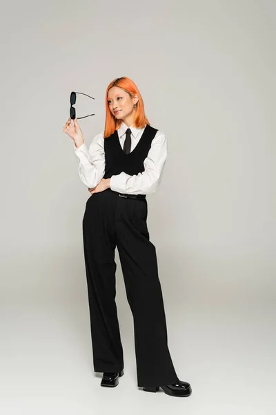 stock image happy emotion, full length of asian woman with dyed red hair holding dark sunglasses and looking away on grey background, business casual fashion, white shirt, black tie, vest and pants